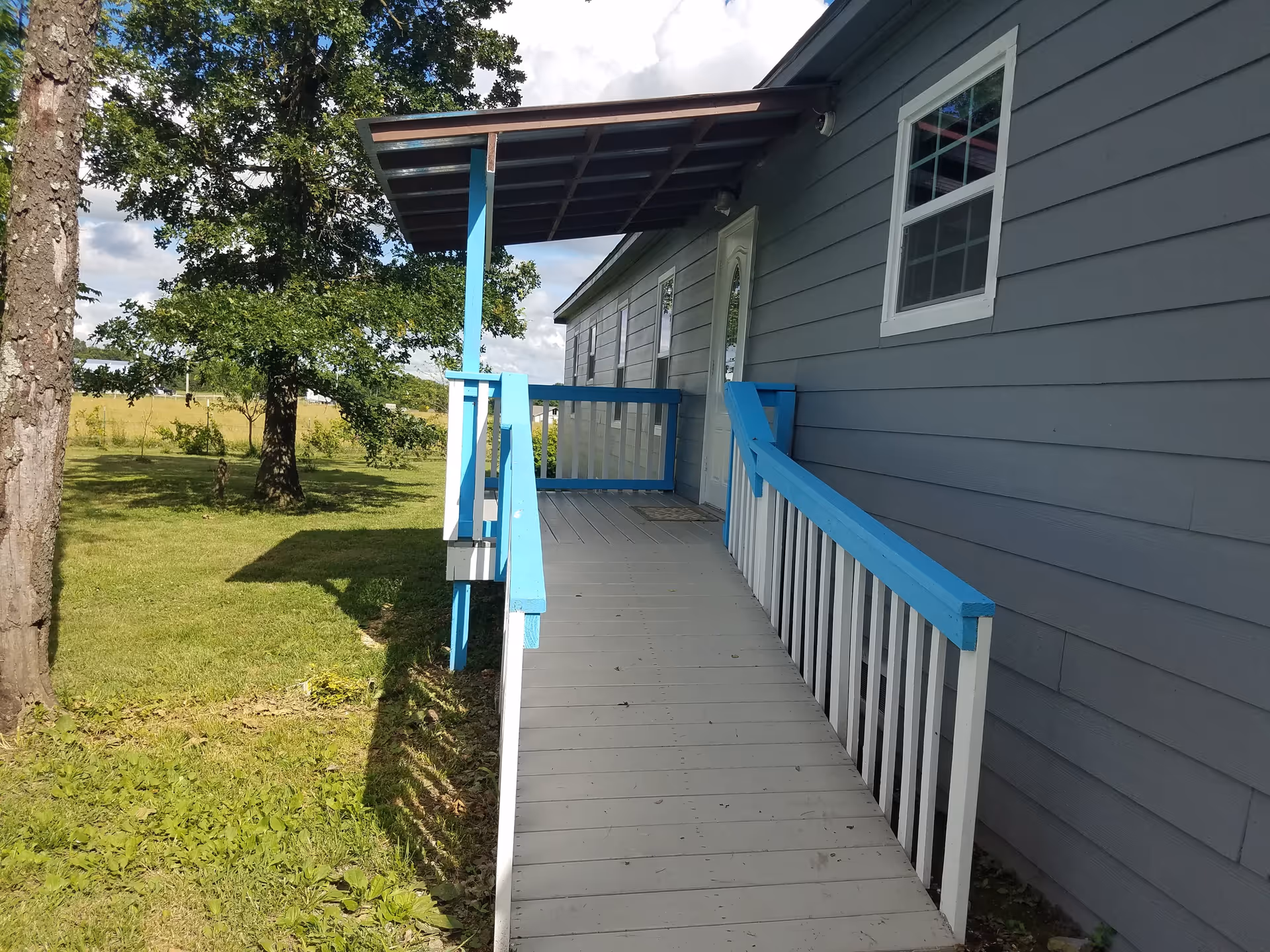 Exterior view of a gray building with a blue and white wheelchair accessible ramp leading to a white door. The ramp has blue handrails and a small roof covering the entrance. There is a grassy area with trees beside the building under a partly cloudy sky.