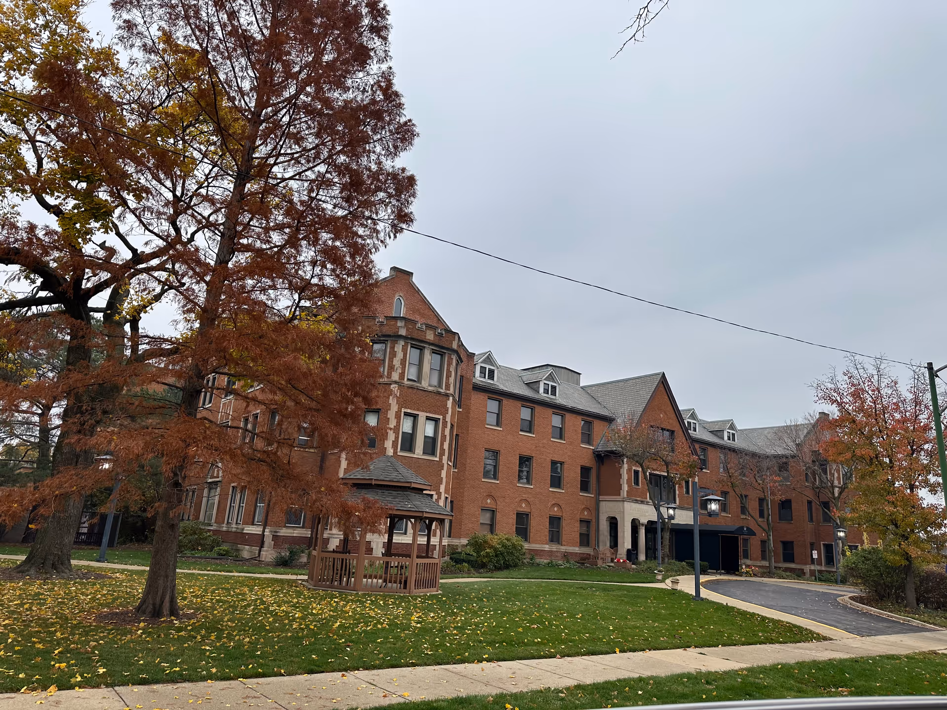Exterior view of a large brick building with multiple windows and a peaked roof, surrounded by trees with autumn foliage and a small wooden gazebo on a grassy lawn. A curved driveway leads to the building entrance under a covered awning.