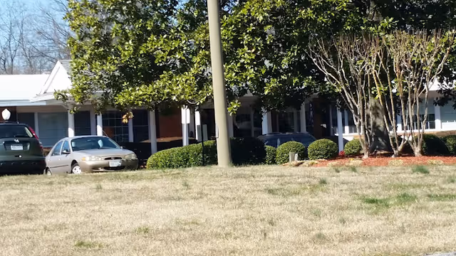 Front view of a single-story rehabilitation and nursing center with a lawn, trees, and parked cars in front.