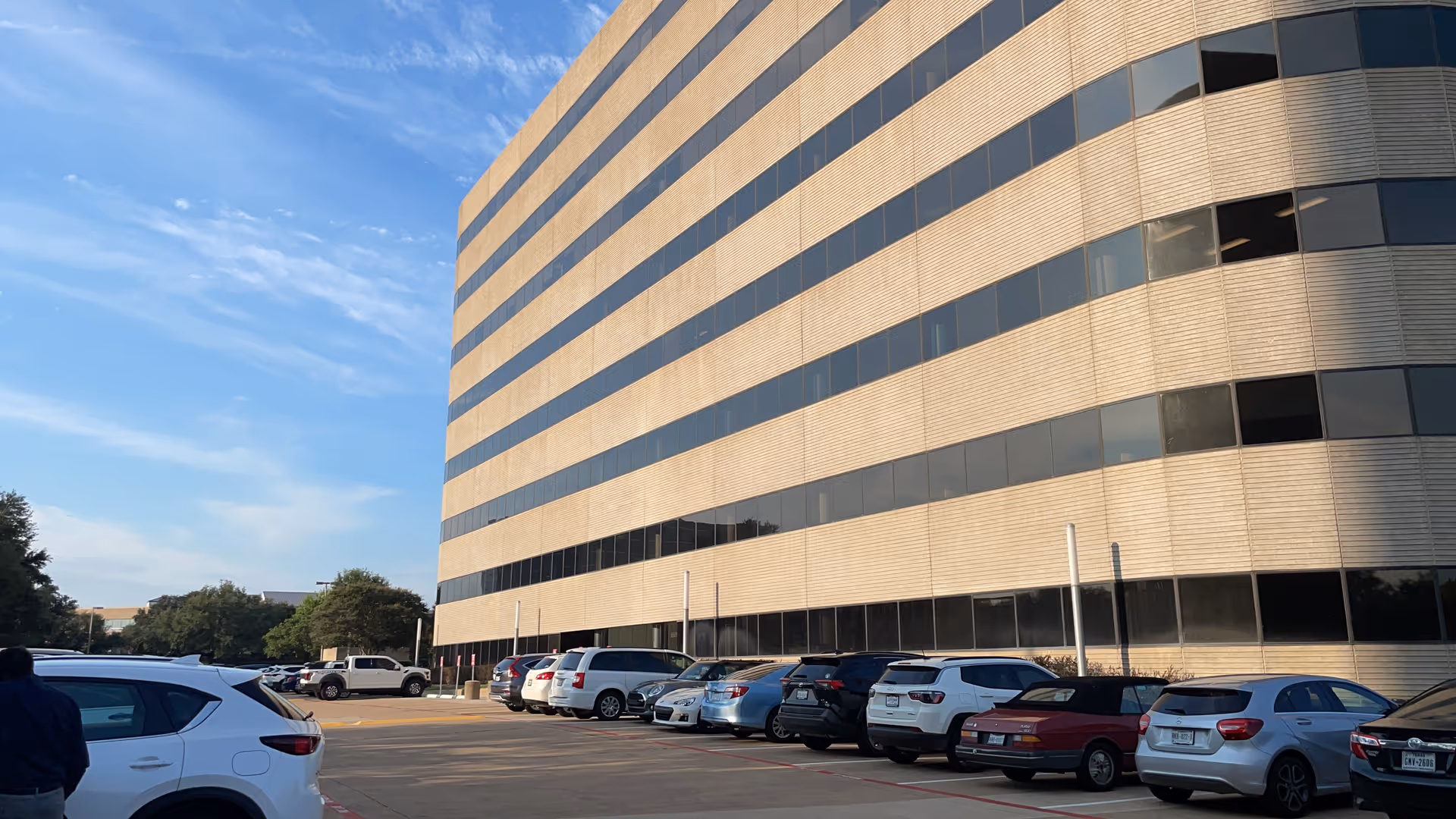 Exterior view of a modern office building with a long curved glass façade and parked cars.