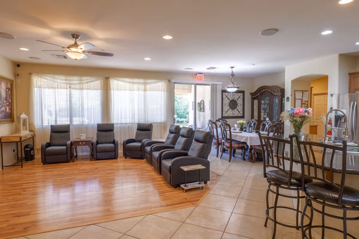 Open communal living area with a row of reclining chairs by large windows, an adjacent dining table, and a kitchen bar with stools.