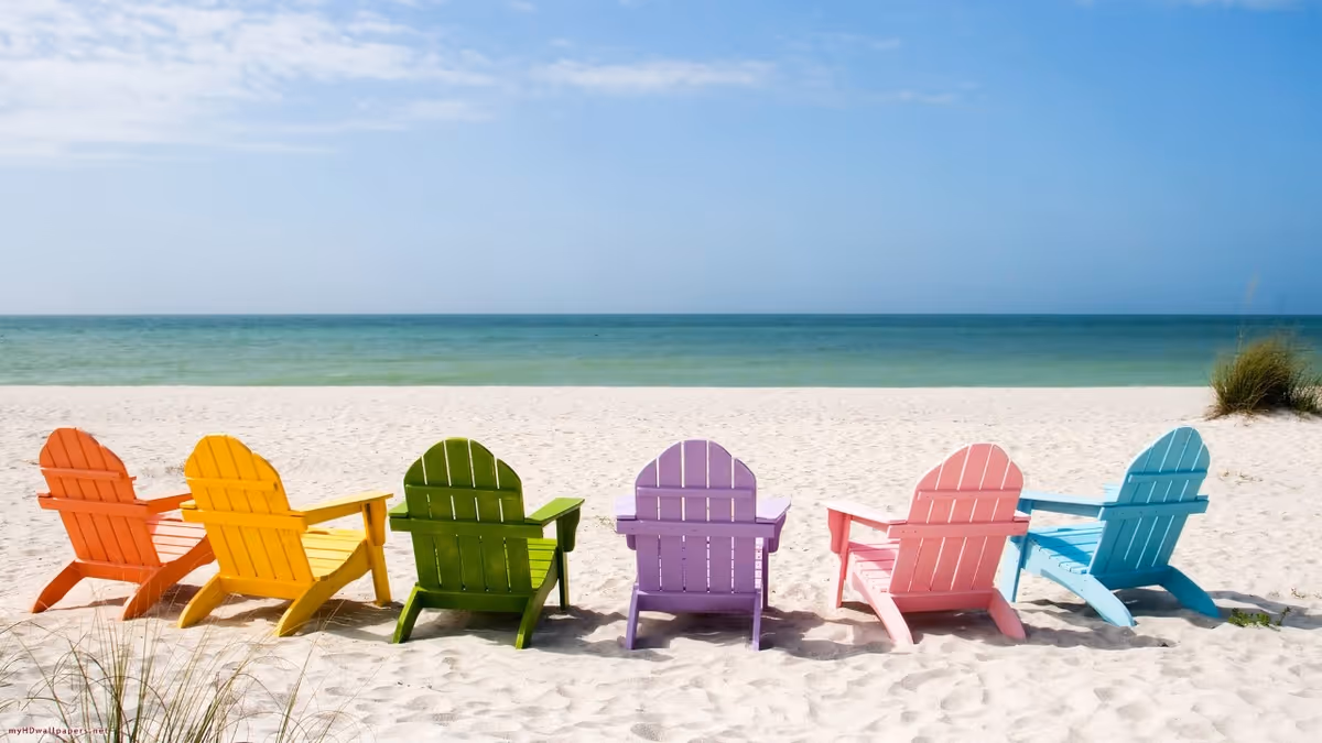 Row of colorful Adirondack chairs on white sand facing a calm ocean.