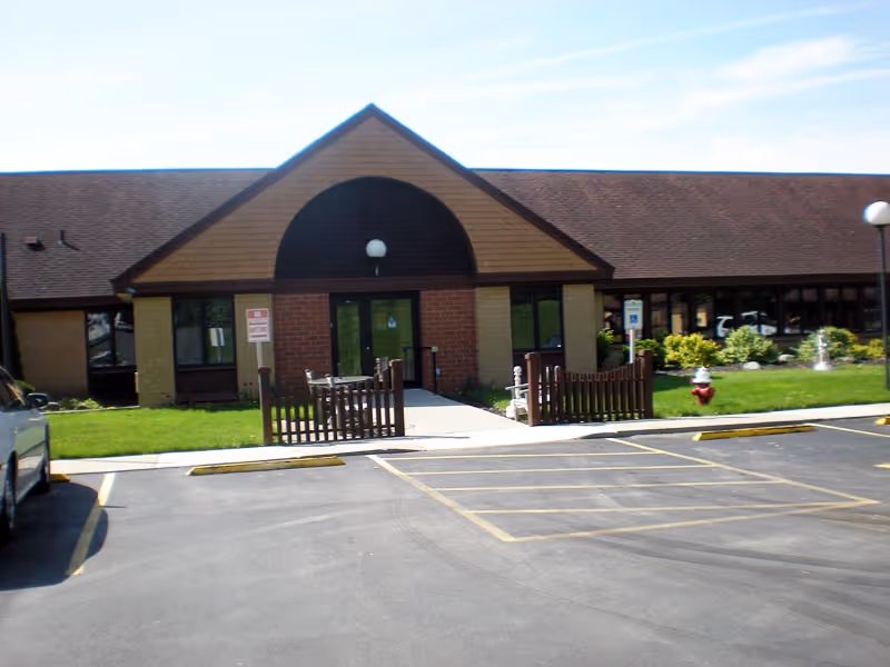 Front entrance of Clinton Crest Manor showing a single-story brick building with a parking lot, ramped walkway, small wooden fence, and landscaped lawn.