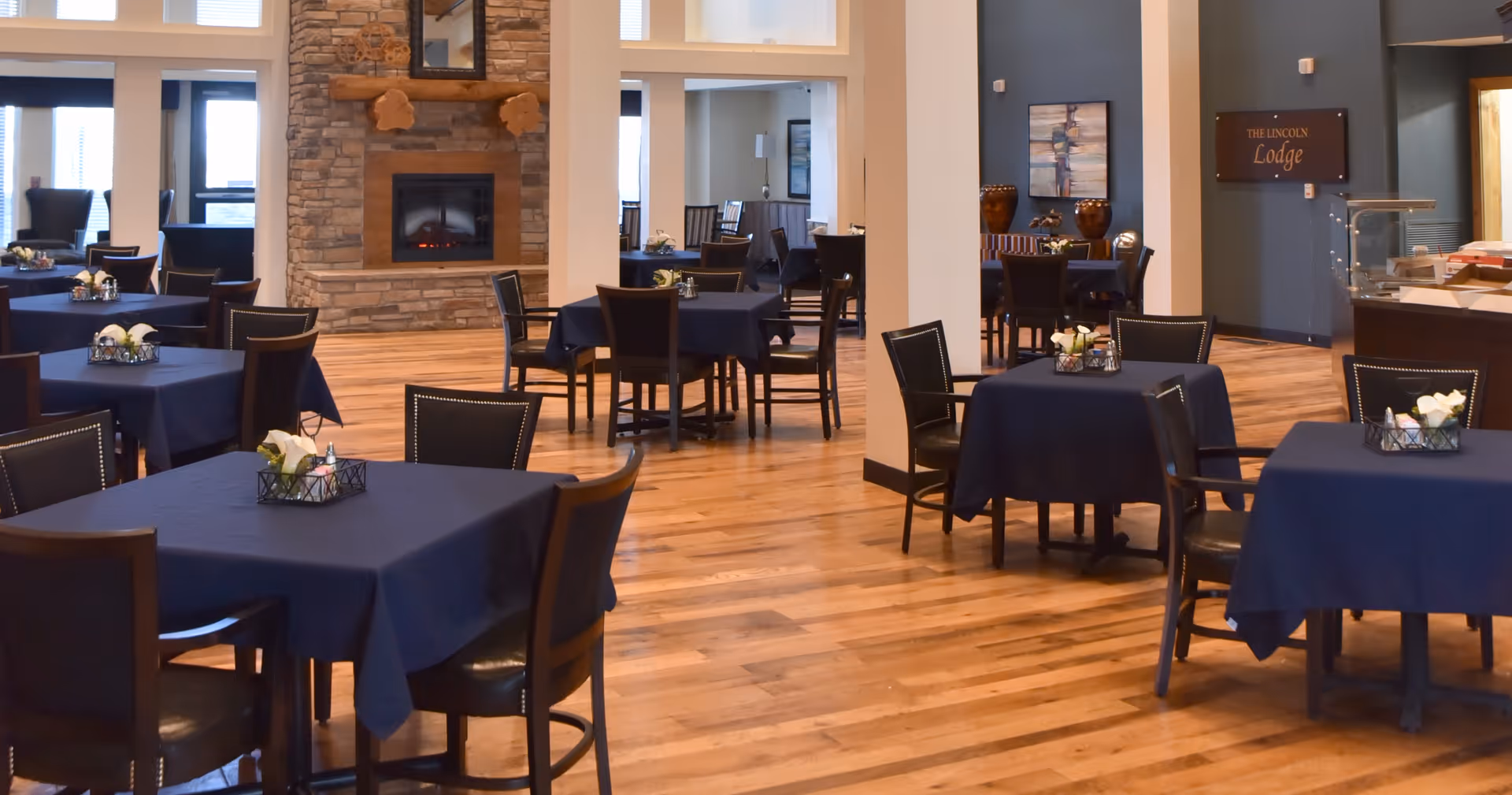A spacious dining area with multiple tables covered in dark blue tablecloths, each set with a small floral centerpiece. The room features wooden flooring, a stone fireplace with a wooden mantle, and large windows allowing natural light. Chairs are arranged around the tables, and there is a sign on the wall that reads 'The Lincoln Lodge'.