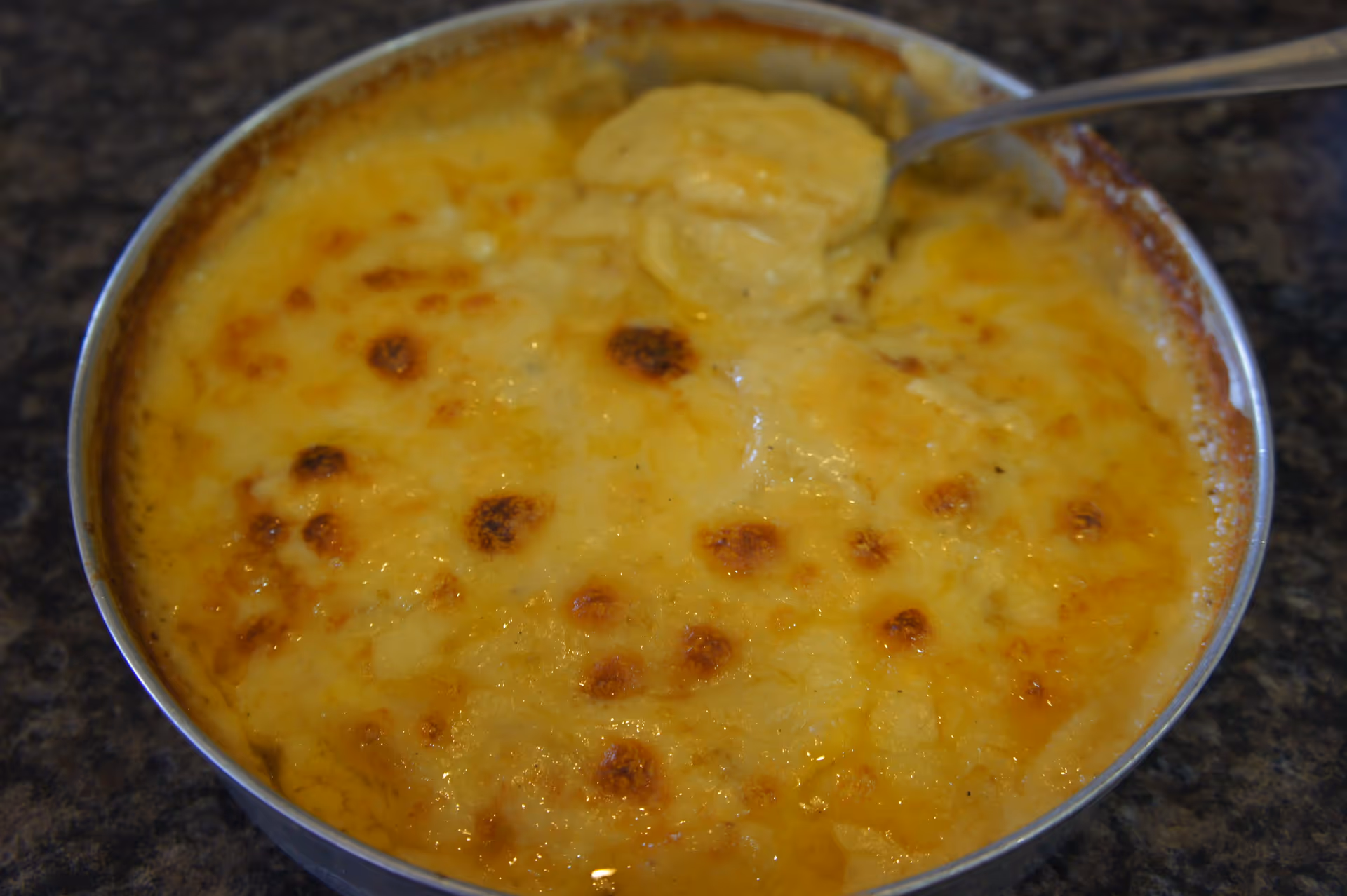 A close-up view of a round dish filled with baked scalloped potatoes topped with melted and browned cheese, with a spoon scooping some of the potatoes.