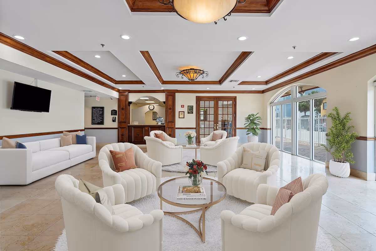 A bright and spacious living room area in a senior living facility with white cushioned armchairs arranged around a glass coffee table holding a vase of flowers and books. The room features a white sofa against the wall with decorative pillows, a wall-mounted TV, wooden ceiling beams, recessed lighting, and large windows letting in natural light. There are potted plants near the windows and a reception desk in the background.