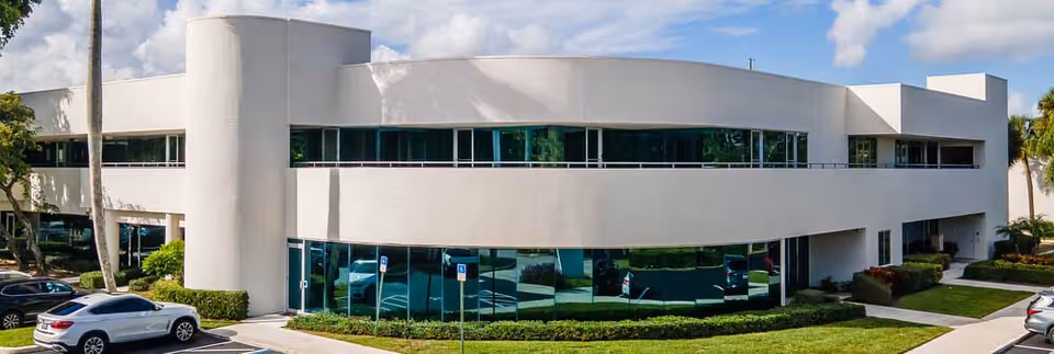 Modern white two-story building with curved facade and large glass windows, parking lot in front.