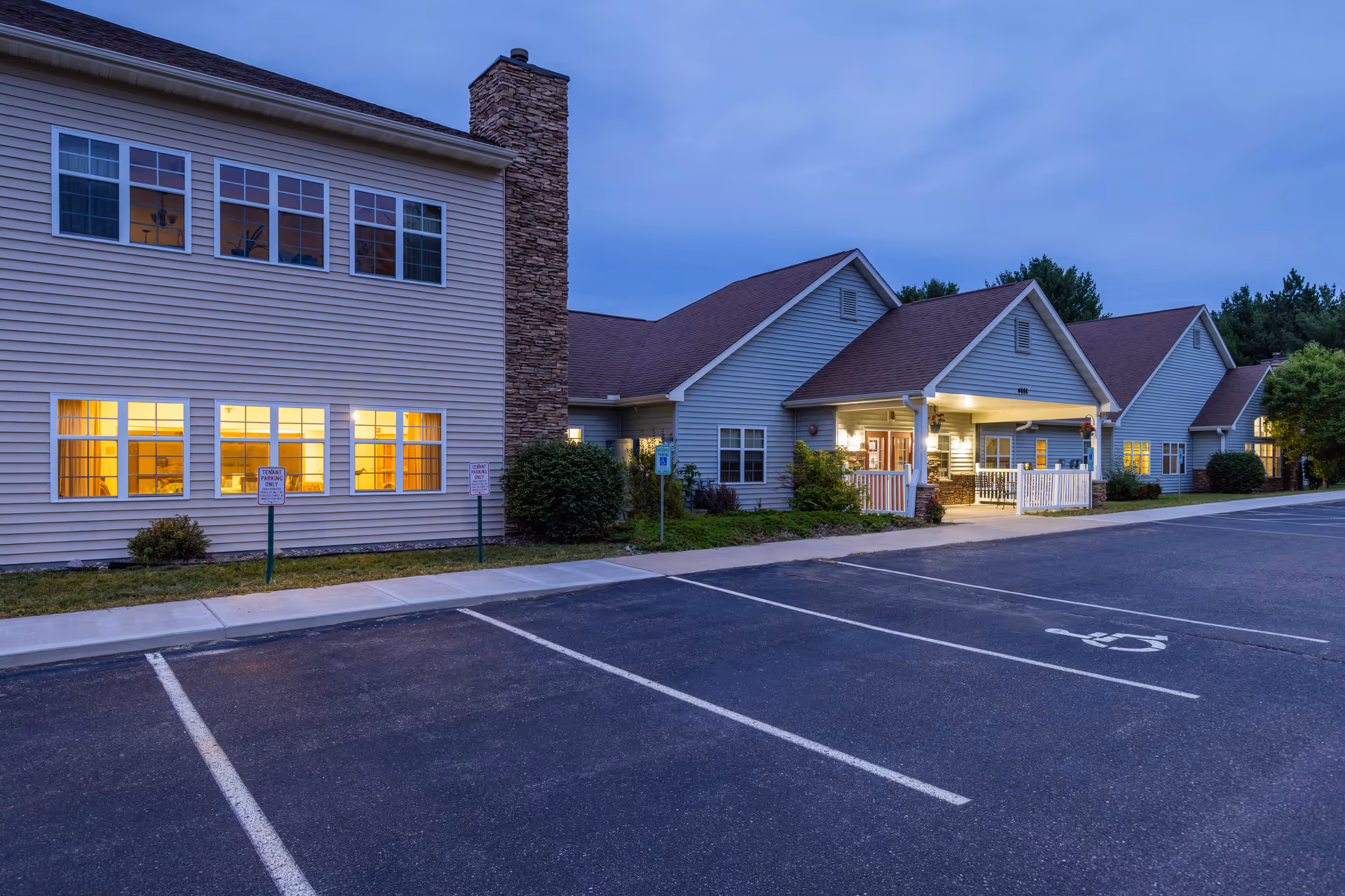 Exterior view of a senior living facility building at dusk with lights on inside. The building has beige siding, a stone chimney, and a covered entrance with white railings. There is a parking lot in front with marked parking spaces, including a handicapped spot.