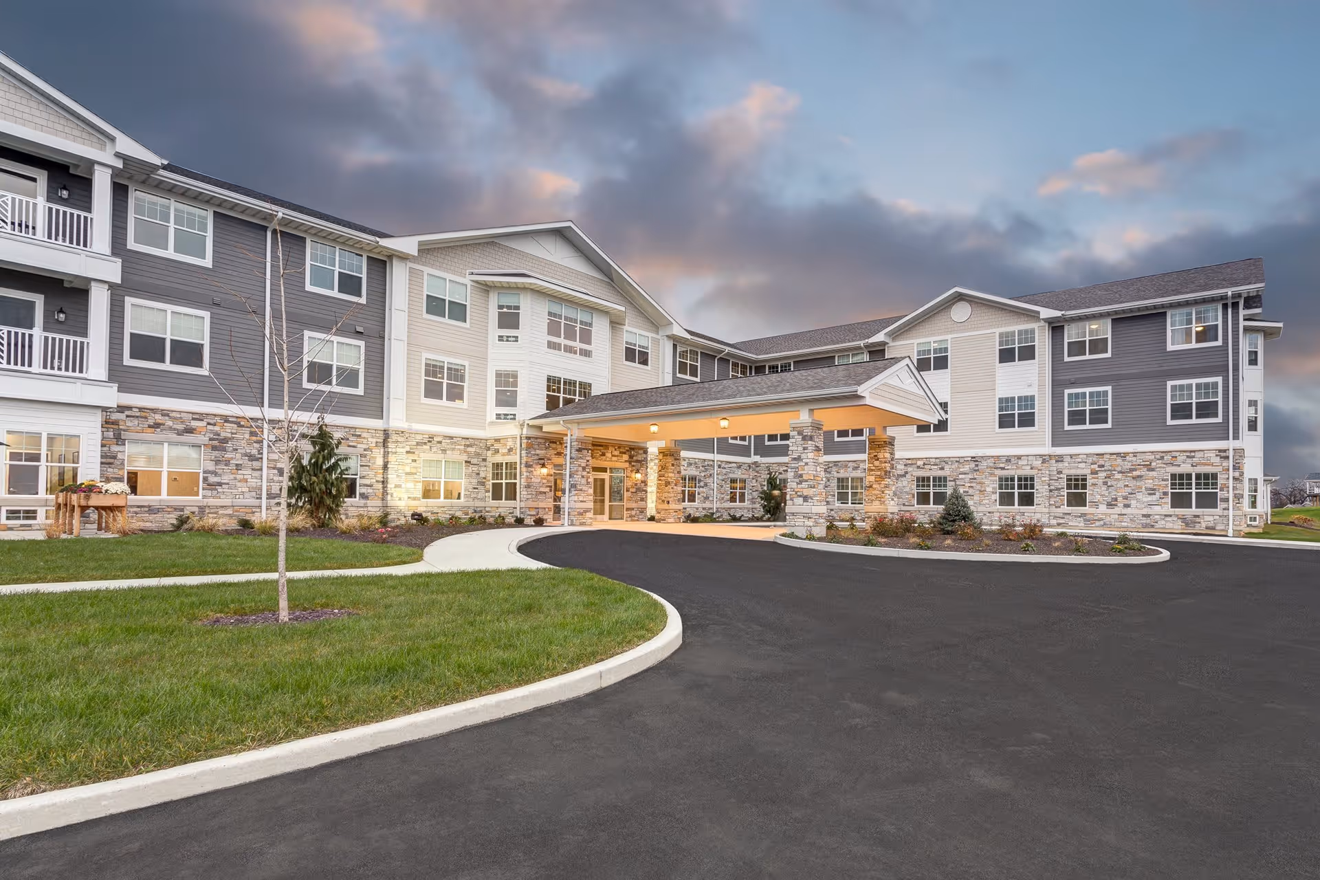 Three-story senior living facility with a covered porte-cochere entrance, landscaped lawn and circular driveway under a cloudy sky.