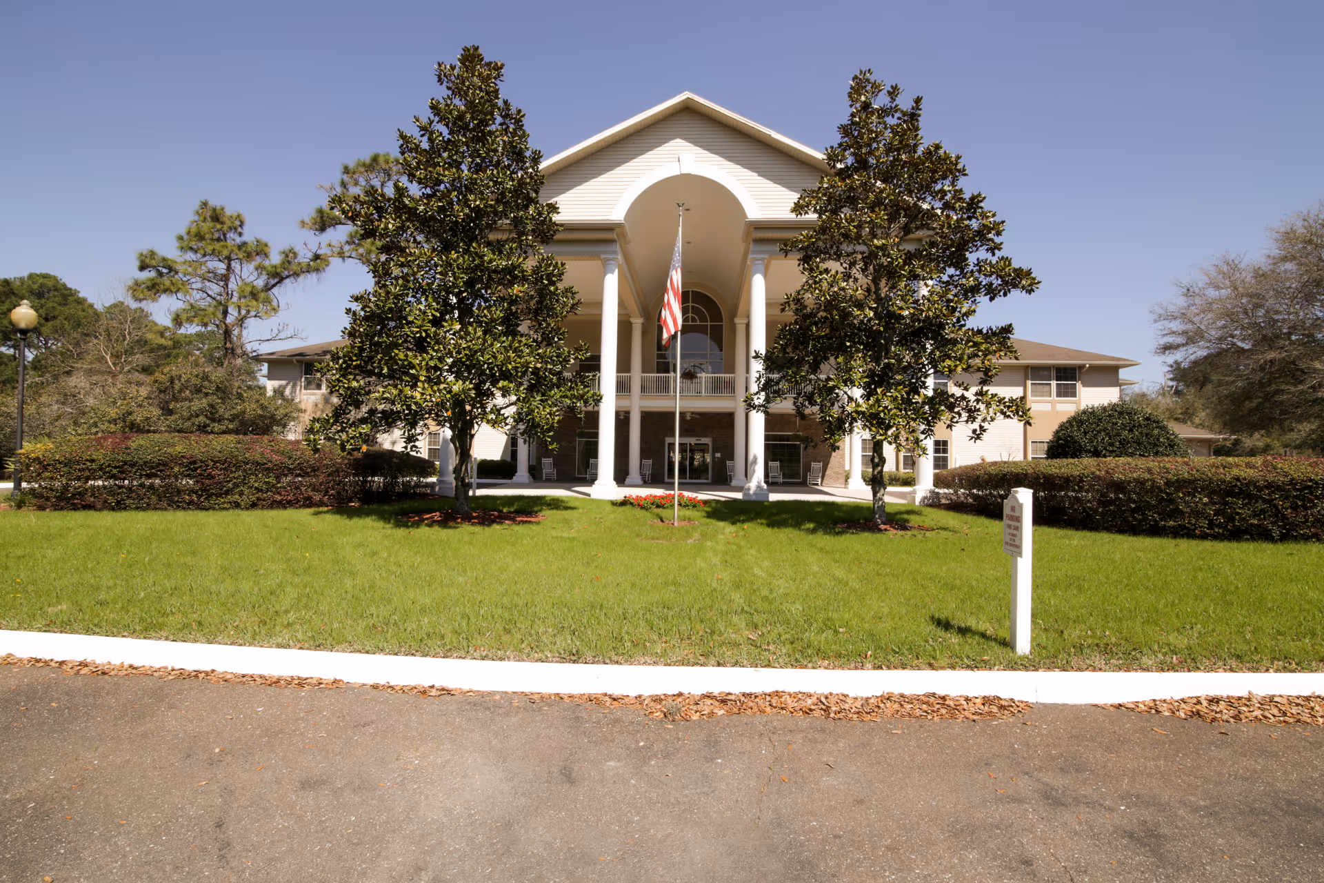 Front exterior of a senior living building with columns, flag, and manicured lawn.