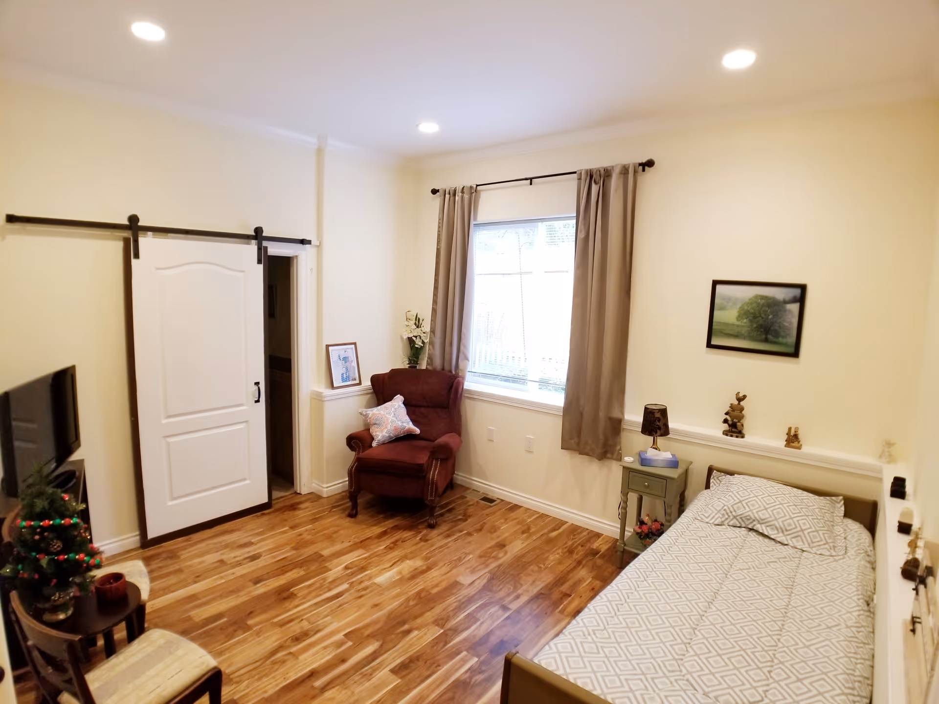 A cozy bedroom with a single bed covered in a patterned gray and white bedspread, a small green nightstand with a lamp and decorative items, a burgundy armchair with a pillow near a window with beige curtains, a sliding barn door, a TV on a stand, and wooden flooring.