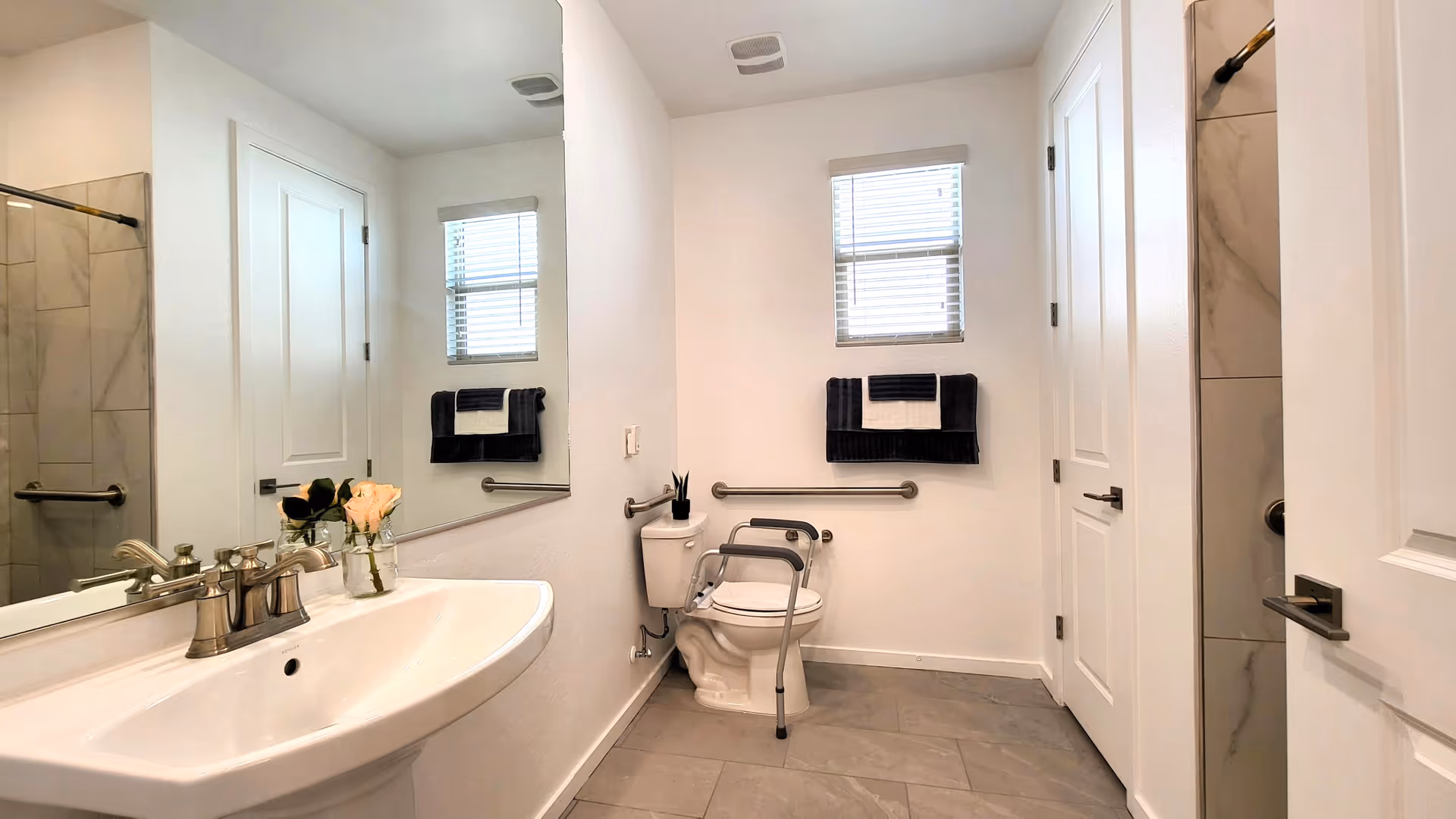 Bright bathroom with sink, mirror, toilet and grab bars, and a tiled shower.