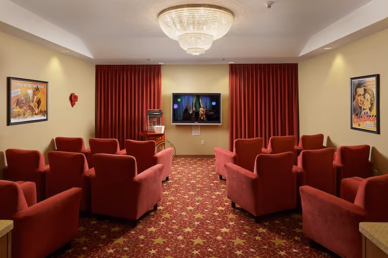 Small screening room with rows of red upholstered armchairs facing a wall-mounted TV, red curtains, a popcorn machine, star-patterned carpet and a chandelier.