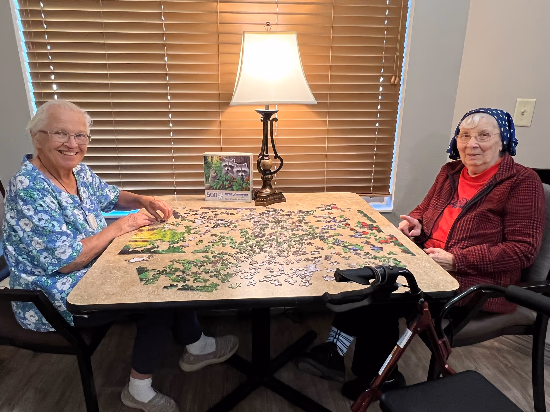 Two elderly women sitting at a table working on a jigsaw puzzle together in a well-lit room with a lamp and closed blinds behind them. One woman is wearing a blue floral top and glasses, smiling at the camera, while the other woman is wearing a red jacket and a blue headscarf. A walker is positioned next to the table.