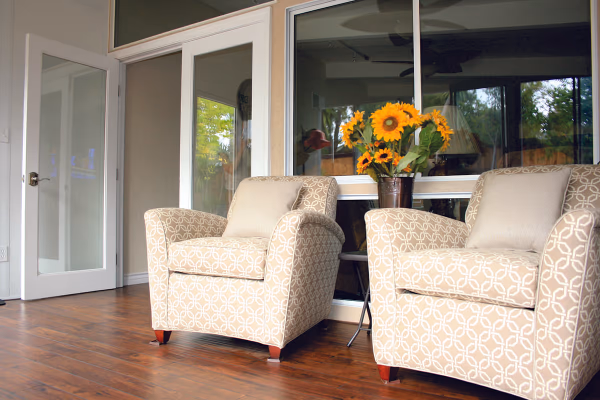 Two patterned armchairs flanking a small table with a vase of sunflowers in a bright room with hardwood floors and glass doors.