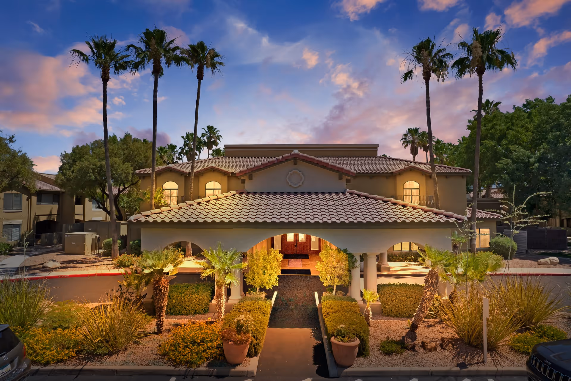 Exterior view of The Country Club of La Cholla building at dusk with a tiled roof, palm trees, and landscaped bushes and plants along the entrance pathway.