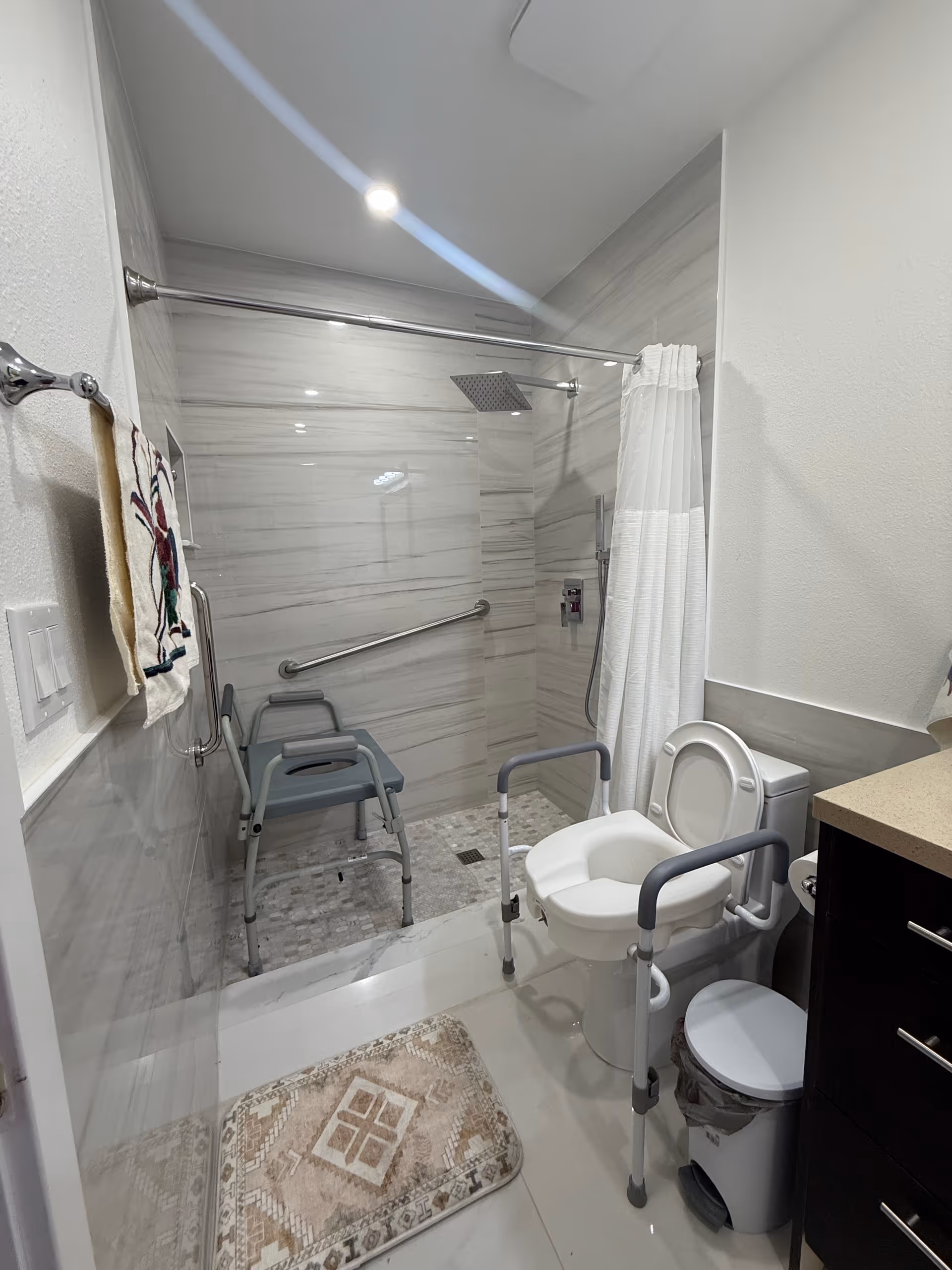 Accessible senior bathroom with toilet, shower chair, grab bars, and curtain.