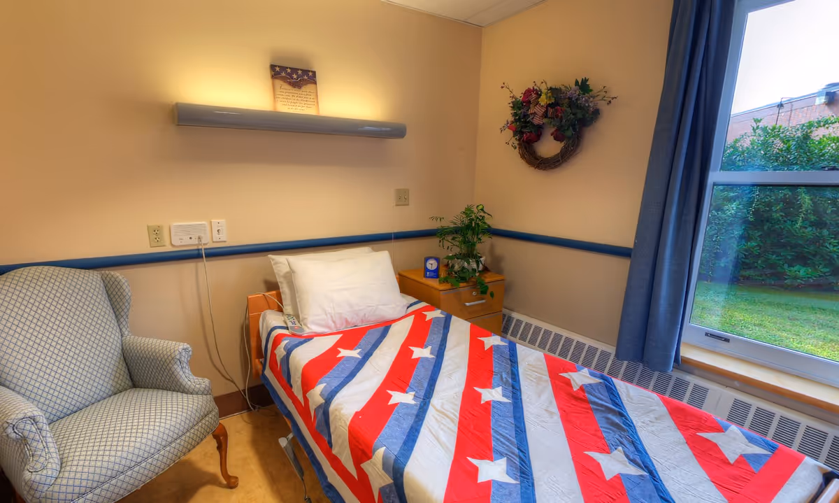Small bedroom in a senior facility with a single bed covered by a red, white, and blue star-spangled quilt, an armchair, nightstand with a plant, wall decor, and a window.