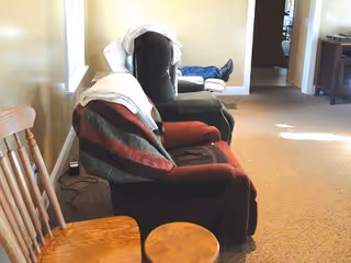 A cozy interior room with two recliner chairs covered with blankets, a wooden chair, and a small round wooden table. The room has beige walls and carpeted floor, with a doorway leading to another room in the background.