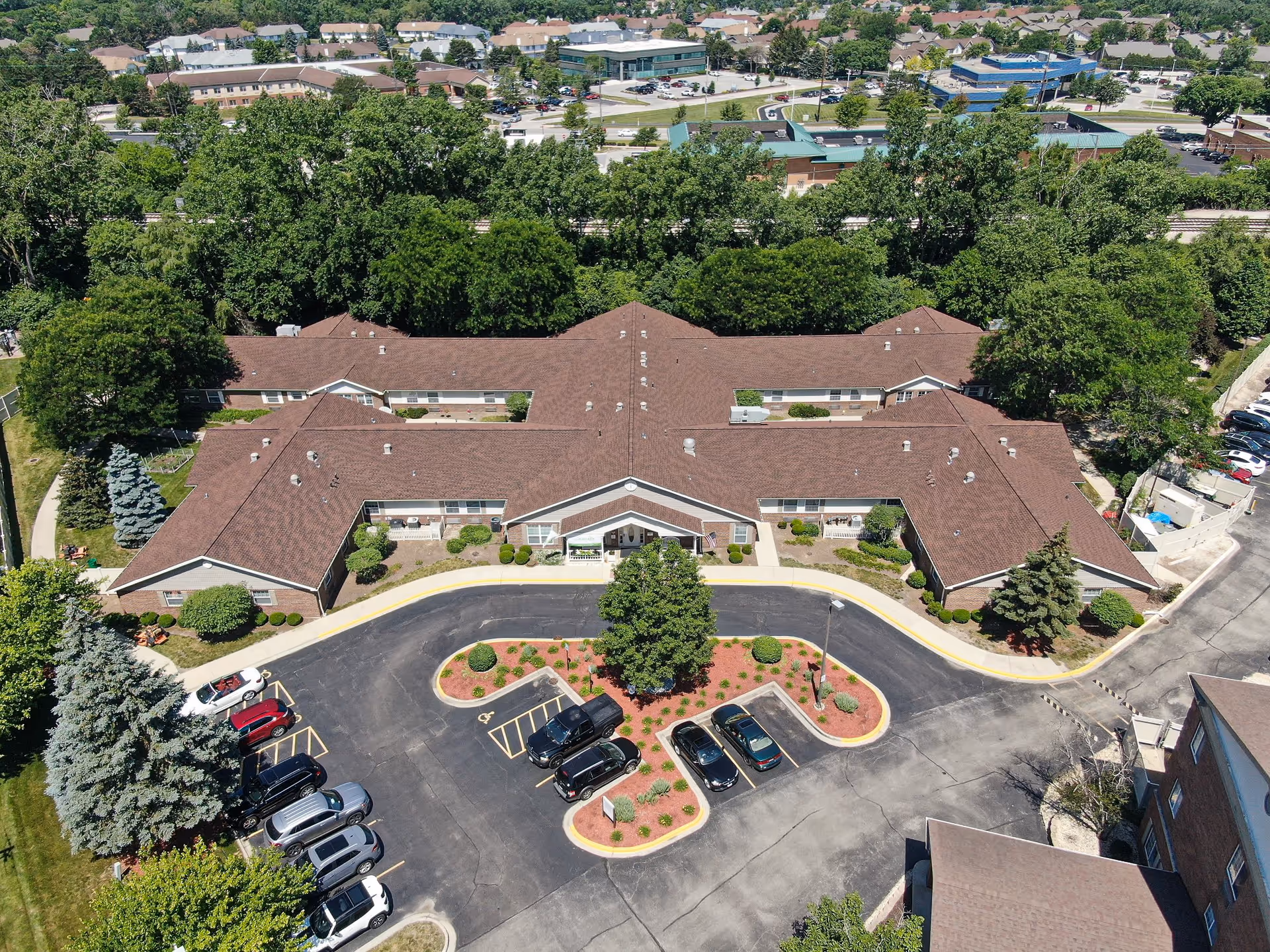 Aerial view of Arden Courts - ProMedica Memory Care Community in Palos Heights, showing a large, single-story building with a brown roof surrounded by trees and landscaping. There is a parking lot with several cars in front of the building and a landscaped island in the center of the parking area.