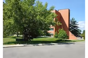 Brick senior living building with trees, green lawn, and a street in front.