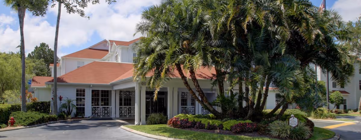 Exterior view of a senior living facility with white buildings and red roofs surrounded by palm trees and landscaped greenery under a partly cloudy sky.