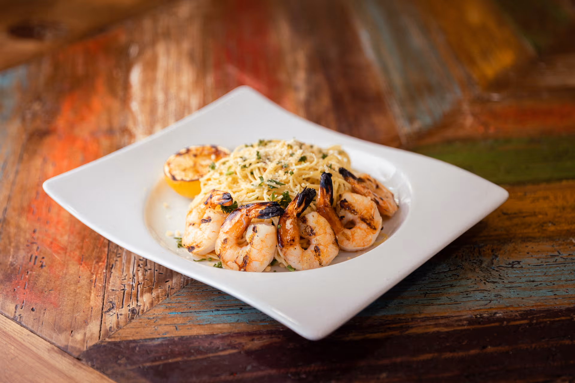 A white square plate with grilled shrimp arranged around a serving of spaghetti pasta garnished with herbs and grated cheese, accompanied by a grilled lemon half, placed on a rustic wooden table.