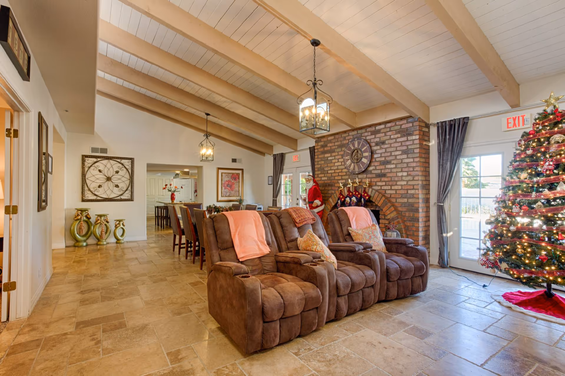 A cozy living room area with three brown recliner chairs adorned with pink blankets and patterned pillows. Behind the chairs is a brick fireplace decorated with a large clock, Christmas stockings, and a Santa Claus figure. To the right, there is a decorated Christmas tree with lights and ornaments near a glass door with an exit sign above. The room has a high ceiling with exposed wooden beams and hanging lantern-style light fixtures. In the background, a dining area with a long table and chairs is visible, along with decorative wall art and vases on the floor.
