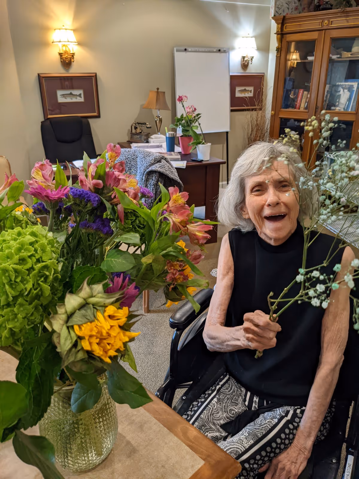 An elderly woman sitting in a wheelchair inside a cozy room, smiling and holding a small bouquet of flowers. In the foreground, there is a vase with a colorful bouquet of flowers on a table. The room has warm lighting, framed fish artwork on the walls, a wooden cabinet with books and decorations, and a desk with a lamp and other items in the background.