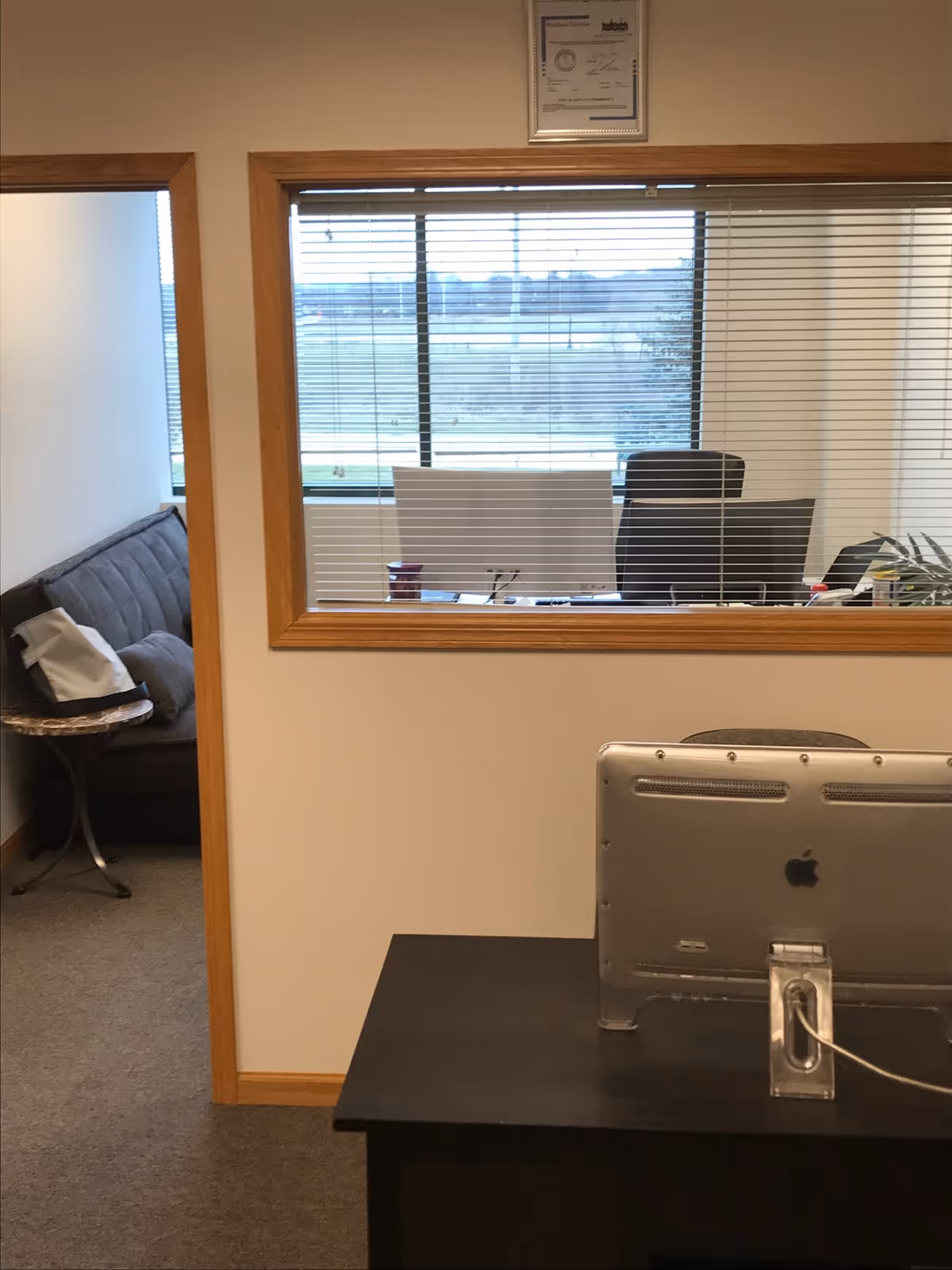 Office interior with desk, computer, blinds, and a seating area beyond the window.