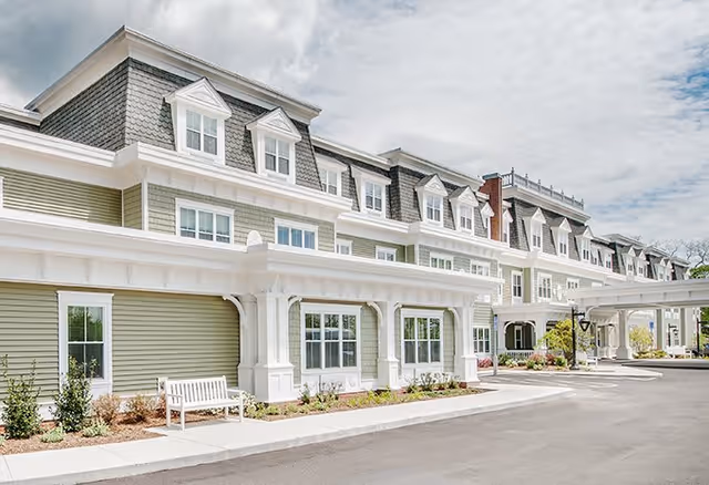 Exterior view of a senior living facility building with light green siding, white trim, multiple windows, and dormer windows on the roof. There is a white bench and some landscaping near the building entrance under a covered driveway.