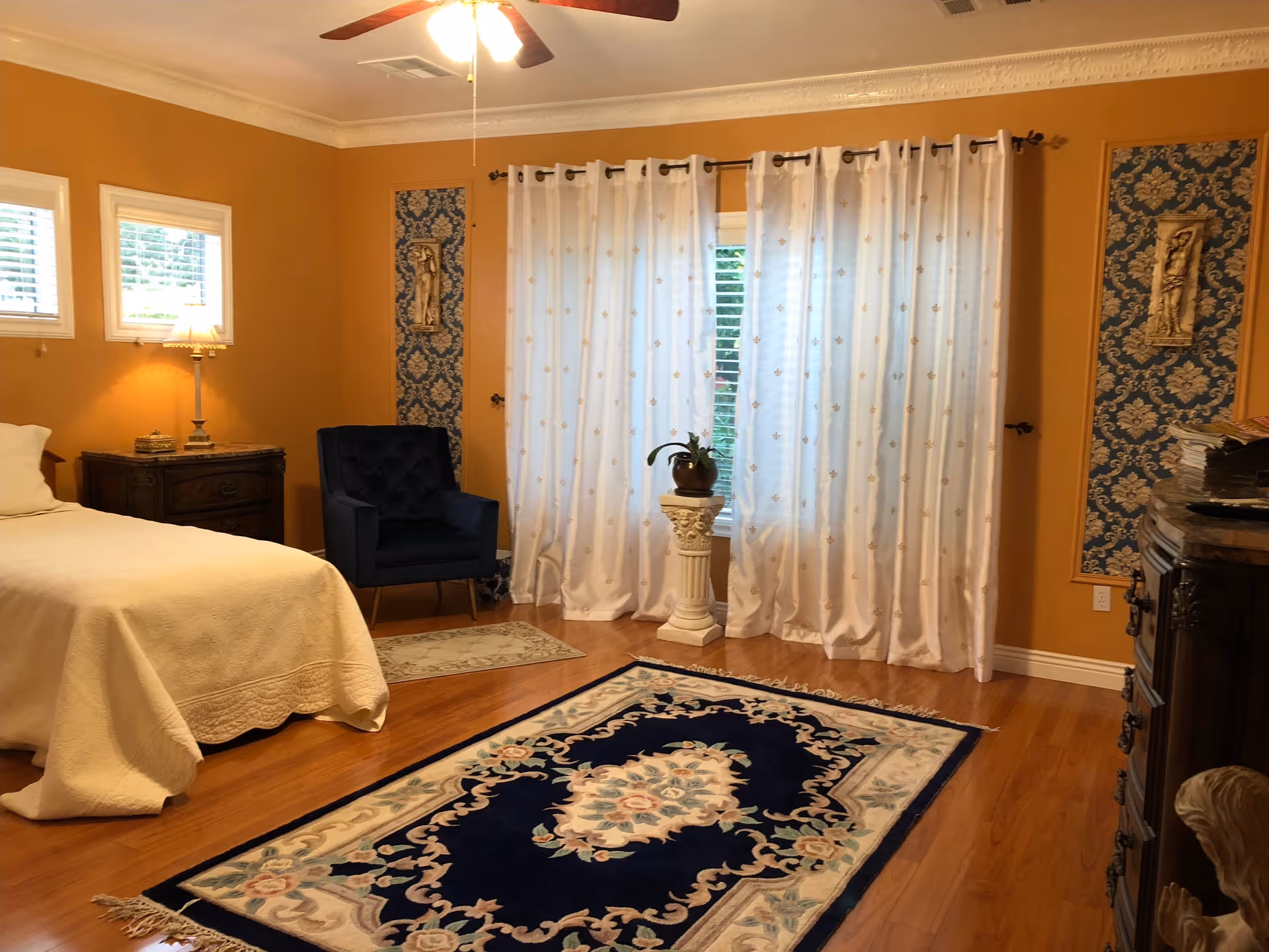 A cozy bedroom with warm orange walls, a bed with a cream-colored quilt, a dark blue armchair, a wooden nightstand with a lamp, white curtains covering a window, decorative wall panels, and a dark blue patterned rug on a wooden floor.