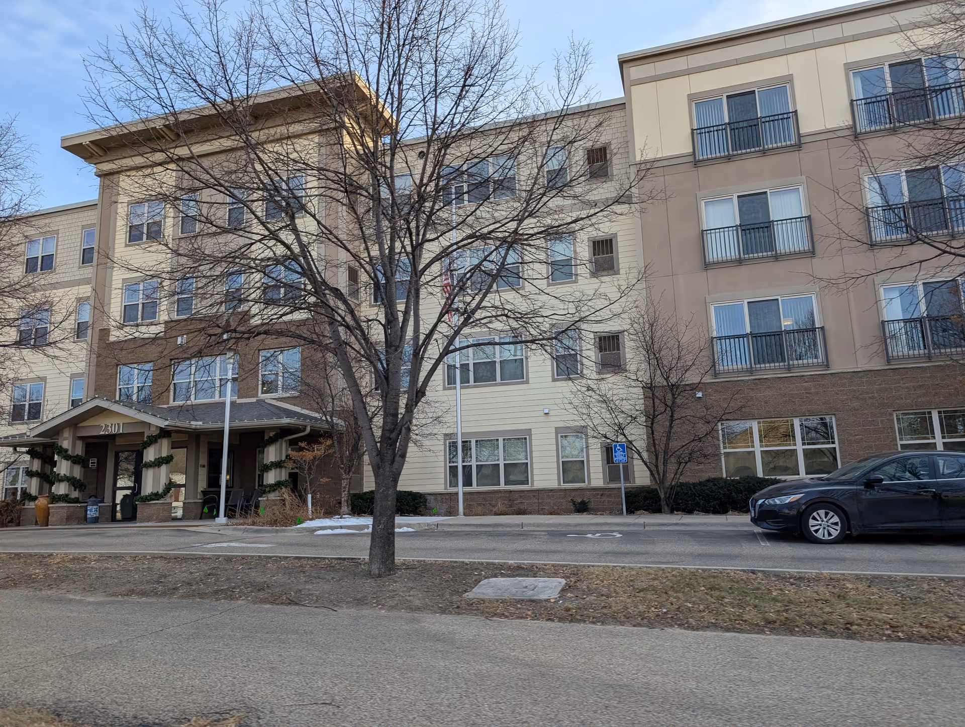 Exterior front view of a multi-story senior living building with a covered entrance, a leafless tree, and a parked car.