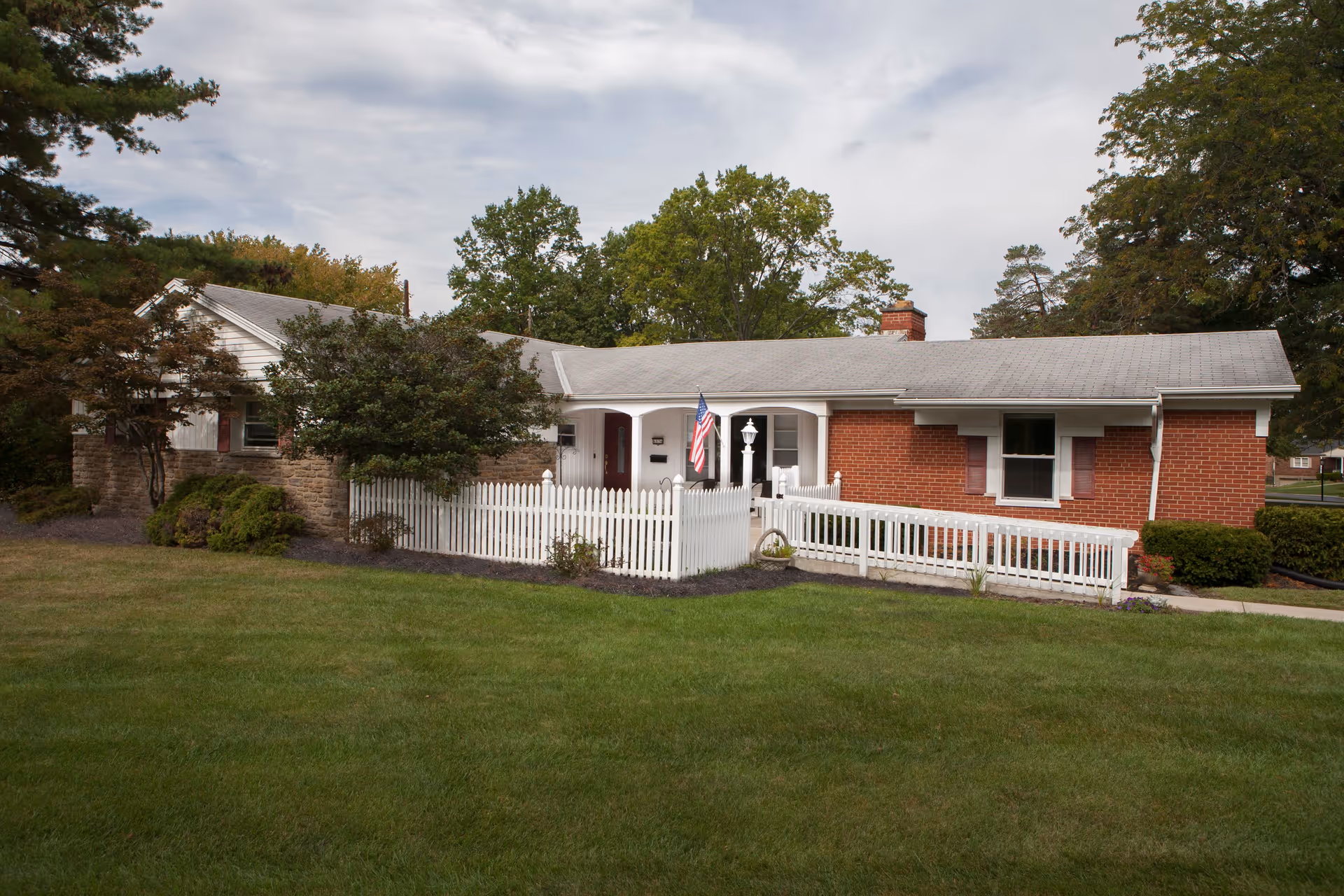 Single-story brick ranch house with a white picket fence, wheelchair ramp, American flag, and a large front lawn.