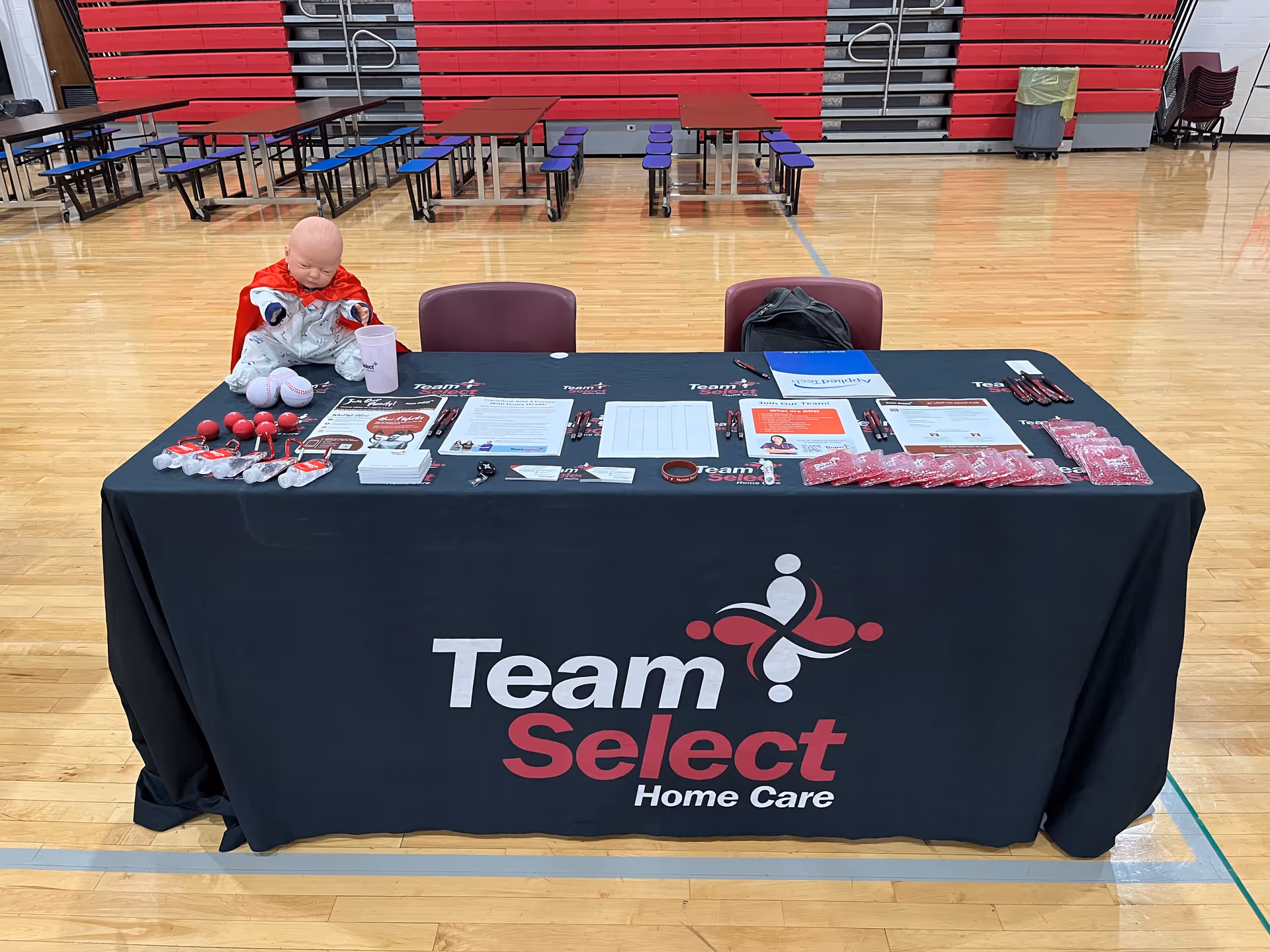 Team Select Home Care information table with brochures and a doll in a gymnasium.