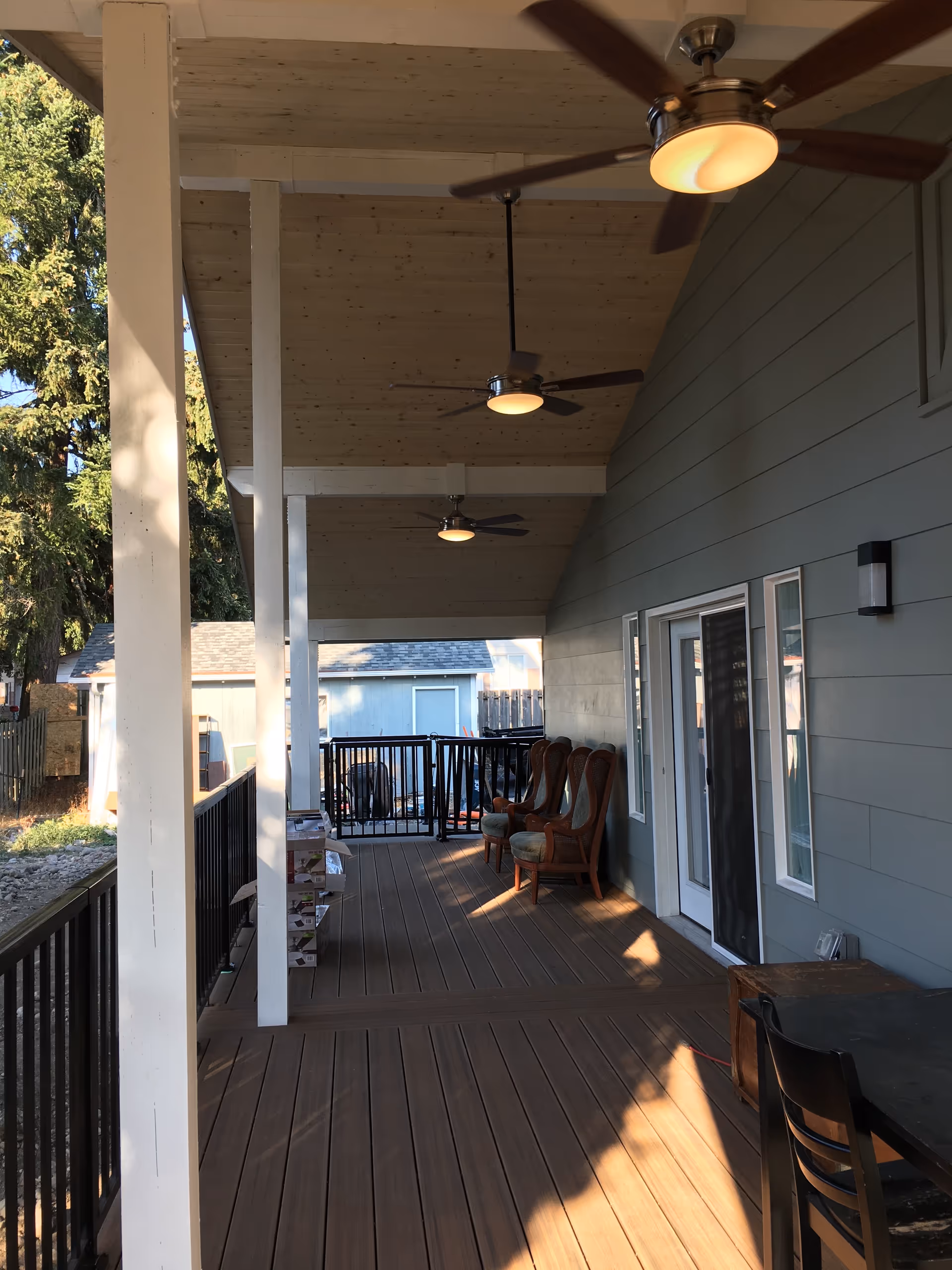 Covered wooden outdoor deck with ceiling fans, chairs, railing, and sliding door to the facility.