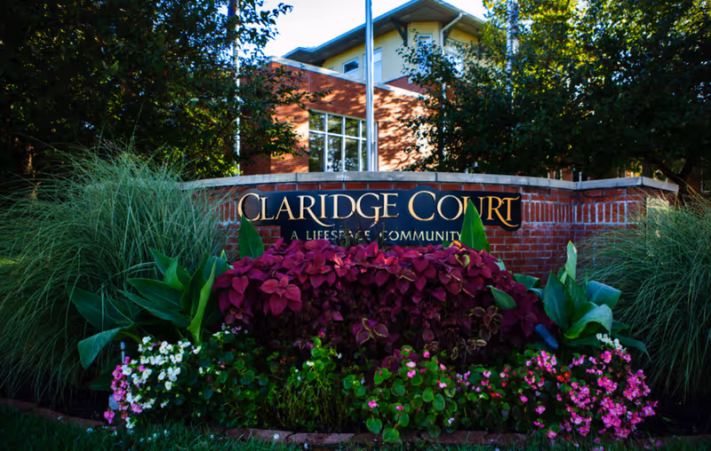 Landscaped brick entrance sign reading Claridge Court with flowers and a multi-story building behind it.