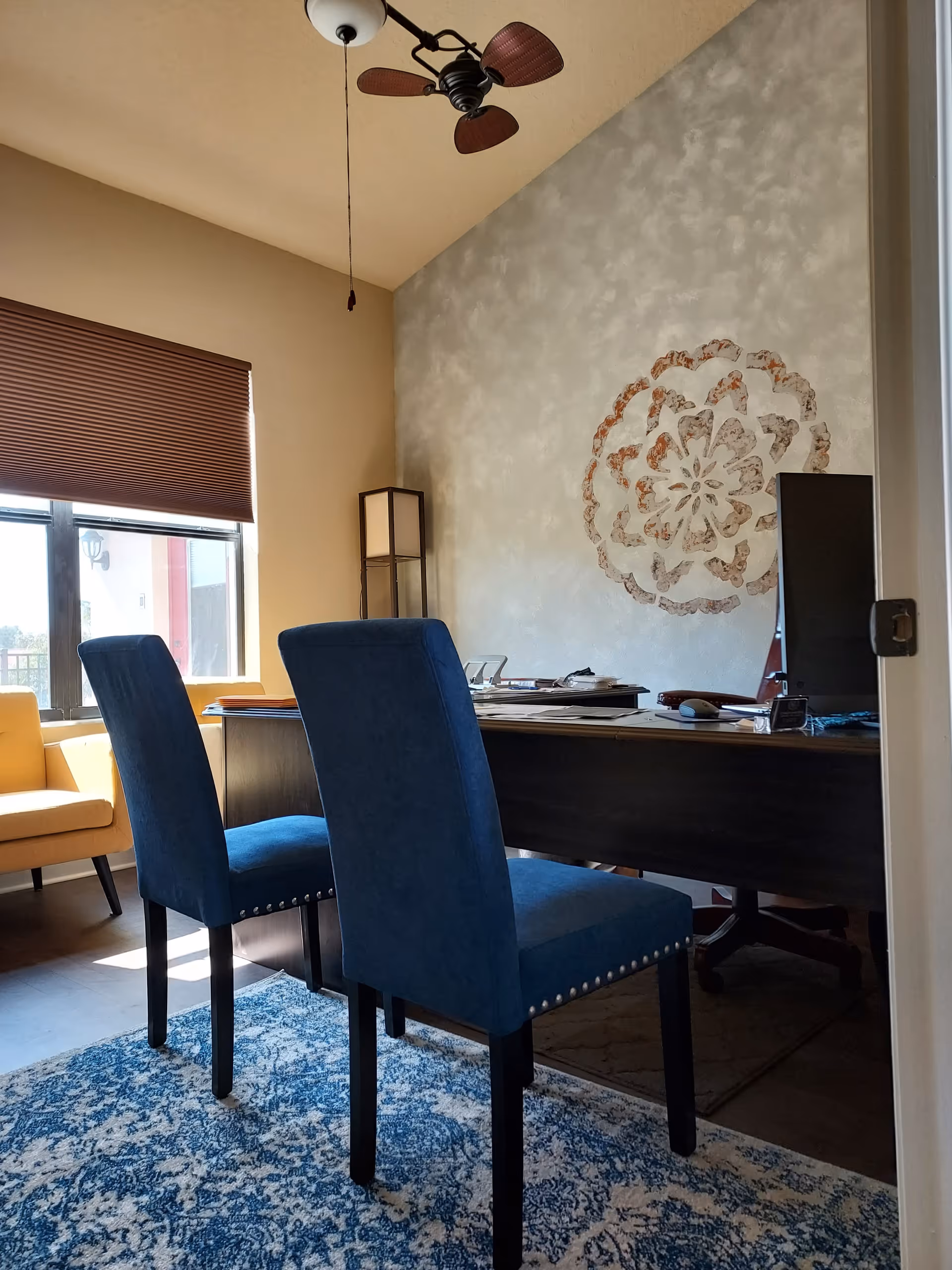 Office-like interior with a desk, blue chairs, yellow chair, blinds, and wall art.