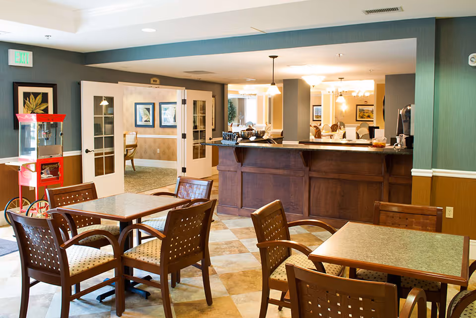 Interior view of a dining area in Southview Assisted Living & Memory Care featuring tables and chairs with cushioned seats, a wooden counter labeled 'Bistro', a red popcorn machine, and double doors leading to another room with additional seating and framed artwork on the walls.