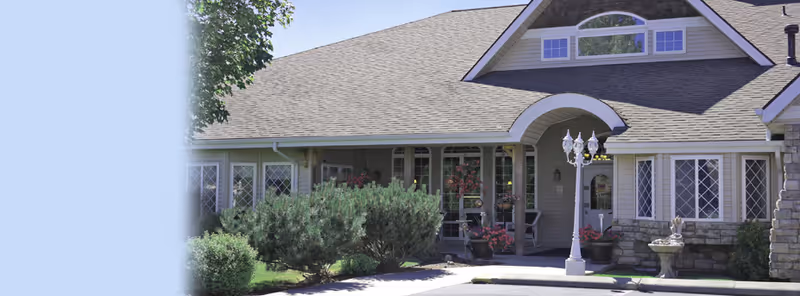 Front exterior view of a residential-style building with a peaked roof, multiple windows with decorative grids, a covered entrance with an arch, a vintage-style street lamp, and landscaping including bushes and flowers.