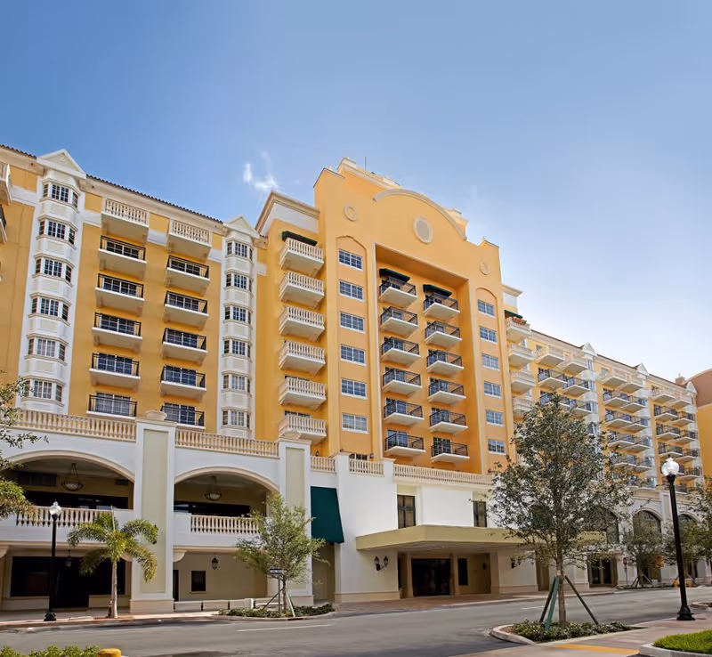 Exterior view of a large multi-story senior living facility building with yellow and white facade, balconies, and arched entrances under a clear blue sky.