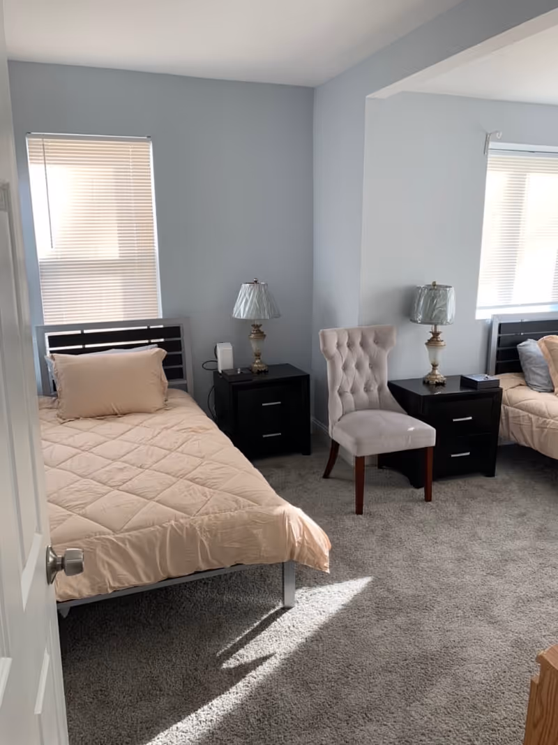 A shared senior living bedroom with two beds, nightstands, lamps, and a tufted chair.