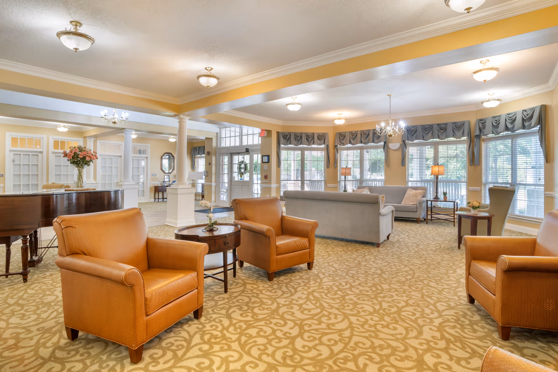 Spacious senior living lobby with orange armchairs, sofas, piano, chandeliers, and large windows.