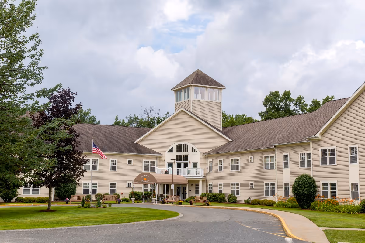 Front exterior view of Benchmark at Orchard Valley, a large beige two-story building with a central tower, surrounded by green trees and a well-maintained lawn with benches and an American flag near the entrance.