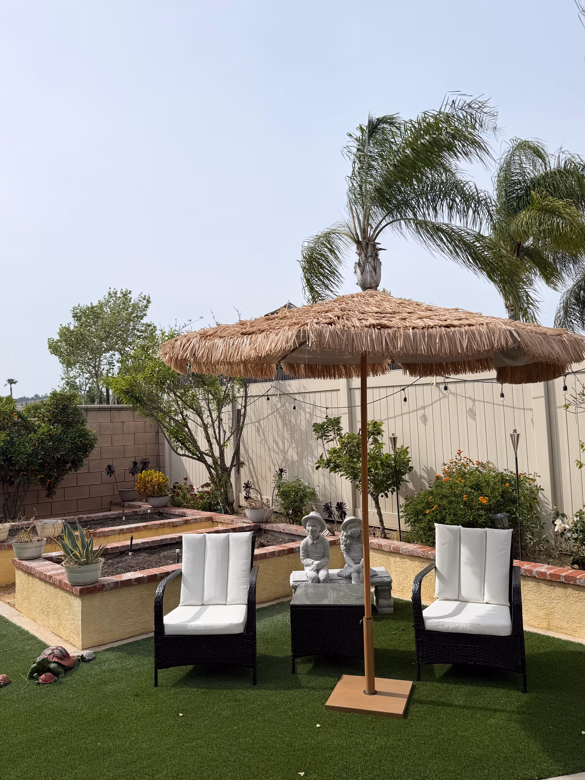 A small outdoor patio with wicker chairs, a thatched umbrella, and garden planters.