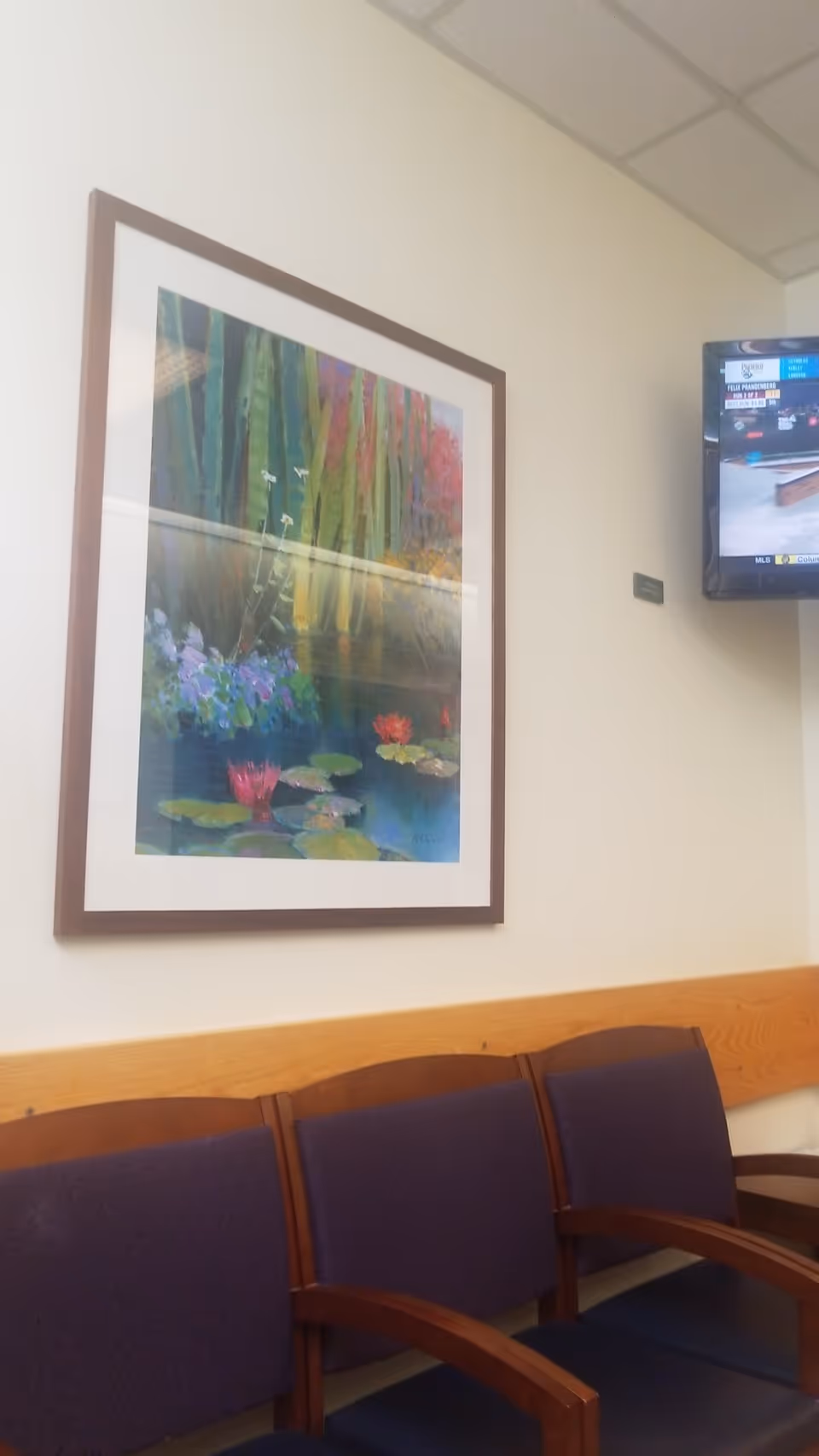 Interior waiting area in a senior living facility with purple chairs, framed art, mounted TV.