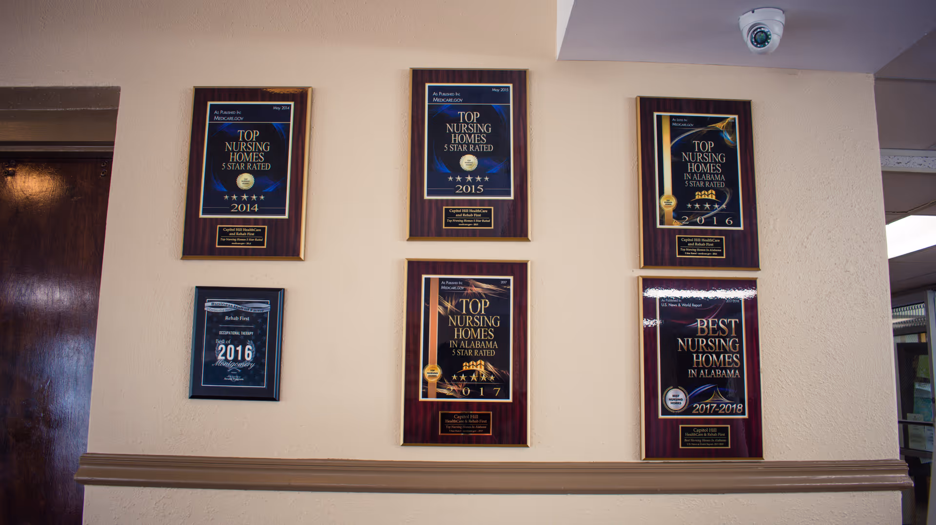 Interior wall displaying six framed nursing home award plaques with a security camera mounted above.