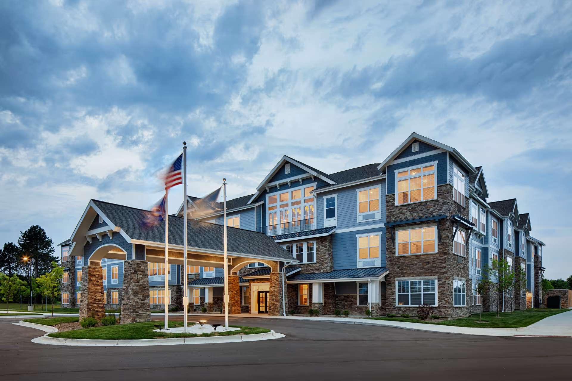 Exterior view of a large, modern senior living facility building with blue siding and stone accents. The building has multiple windows lit from inside and a covered entrance with three flagpoles displaying flags. The sky is partly cloudy during dusk.