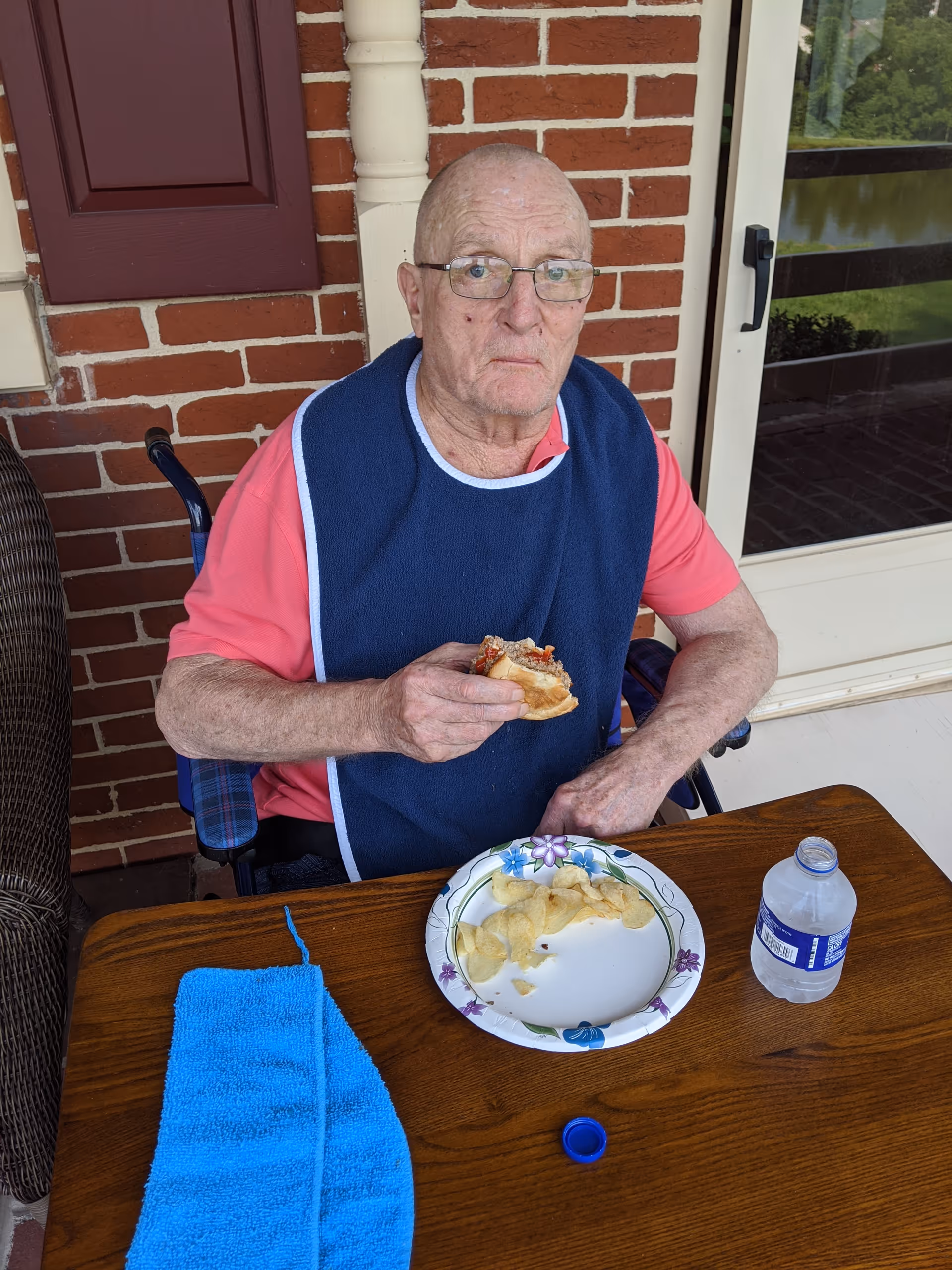 Elderly man sits at a wooden table on a brick porch, eating a sandwich.