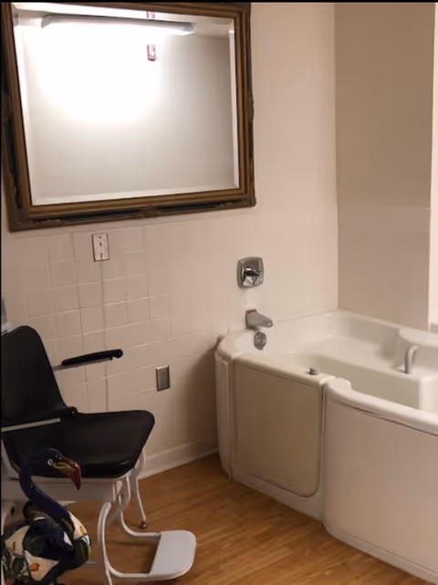A bathroom in a senior living facility featuring a bathtub, mirror, and chair.